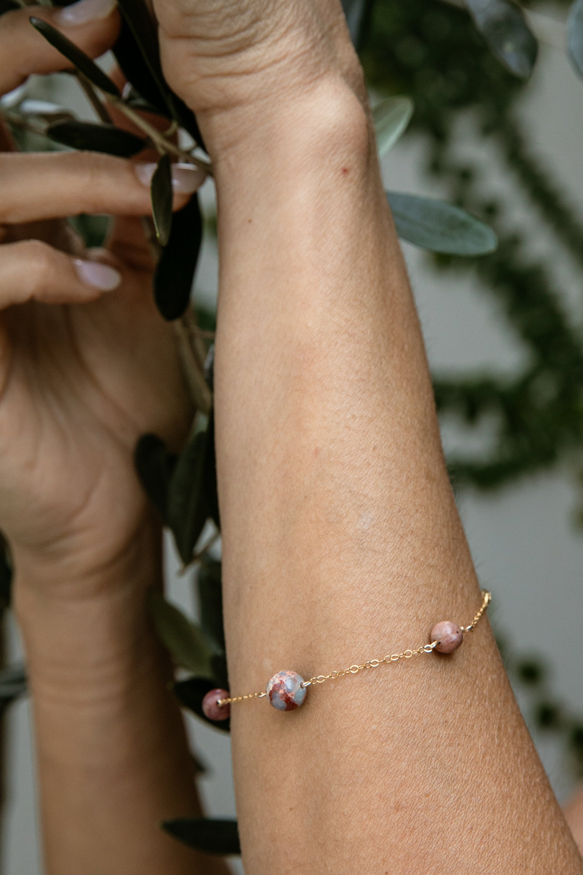 A delicate gold-filled chain bracelet with small pink rhodonite and aqua terra gemstones worn on a person's wrist, shown against a soft-focus background of green leaves.