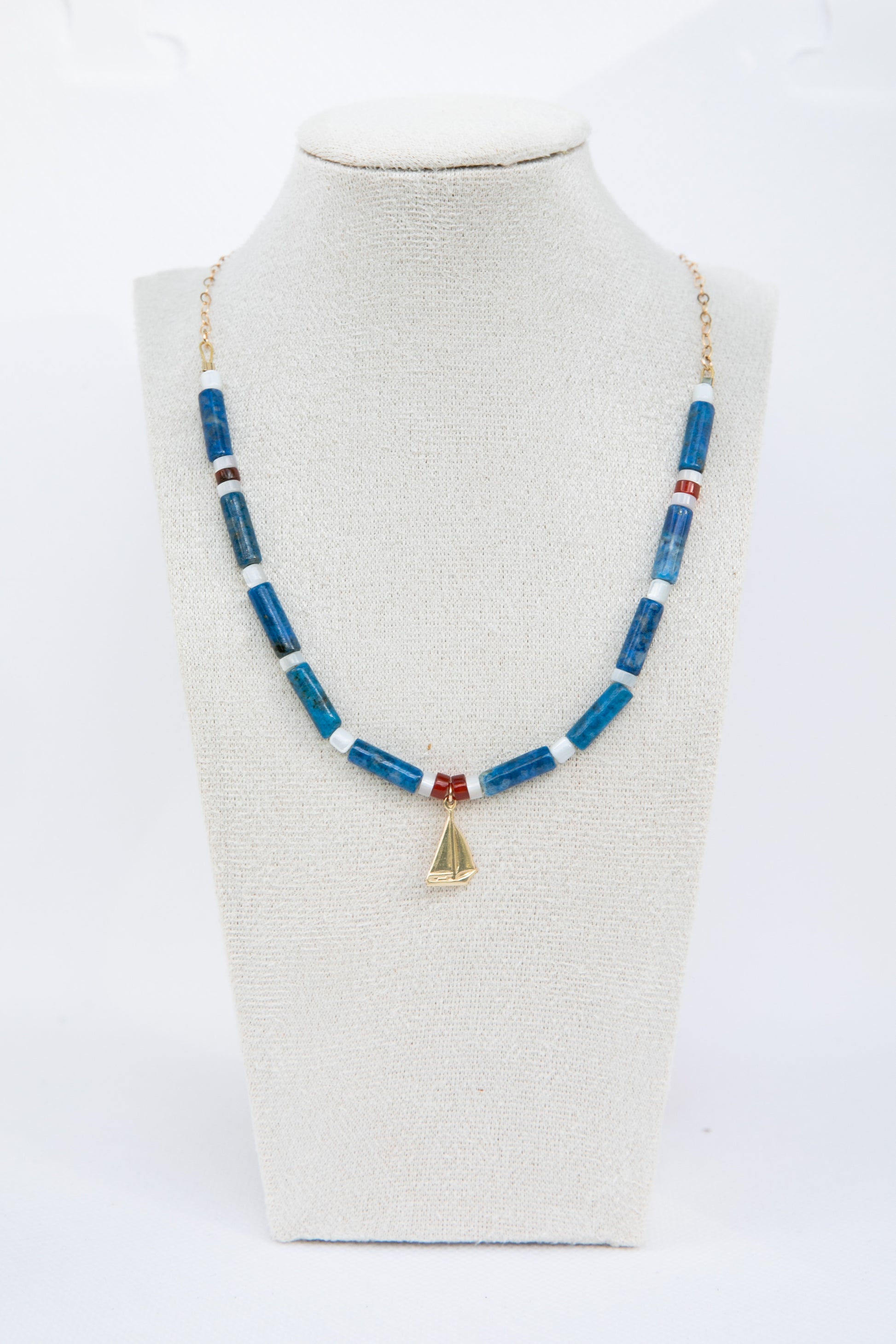 Handcrafted necklace with blue denim lapis, white mother of pearl and read carnelian on a mannequin bust.  The jewelery contains a golf-filled sailing boat charm as an homage to the South of France and sailing culture. 