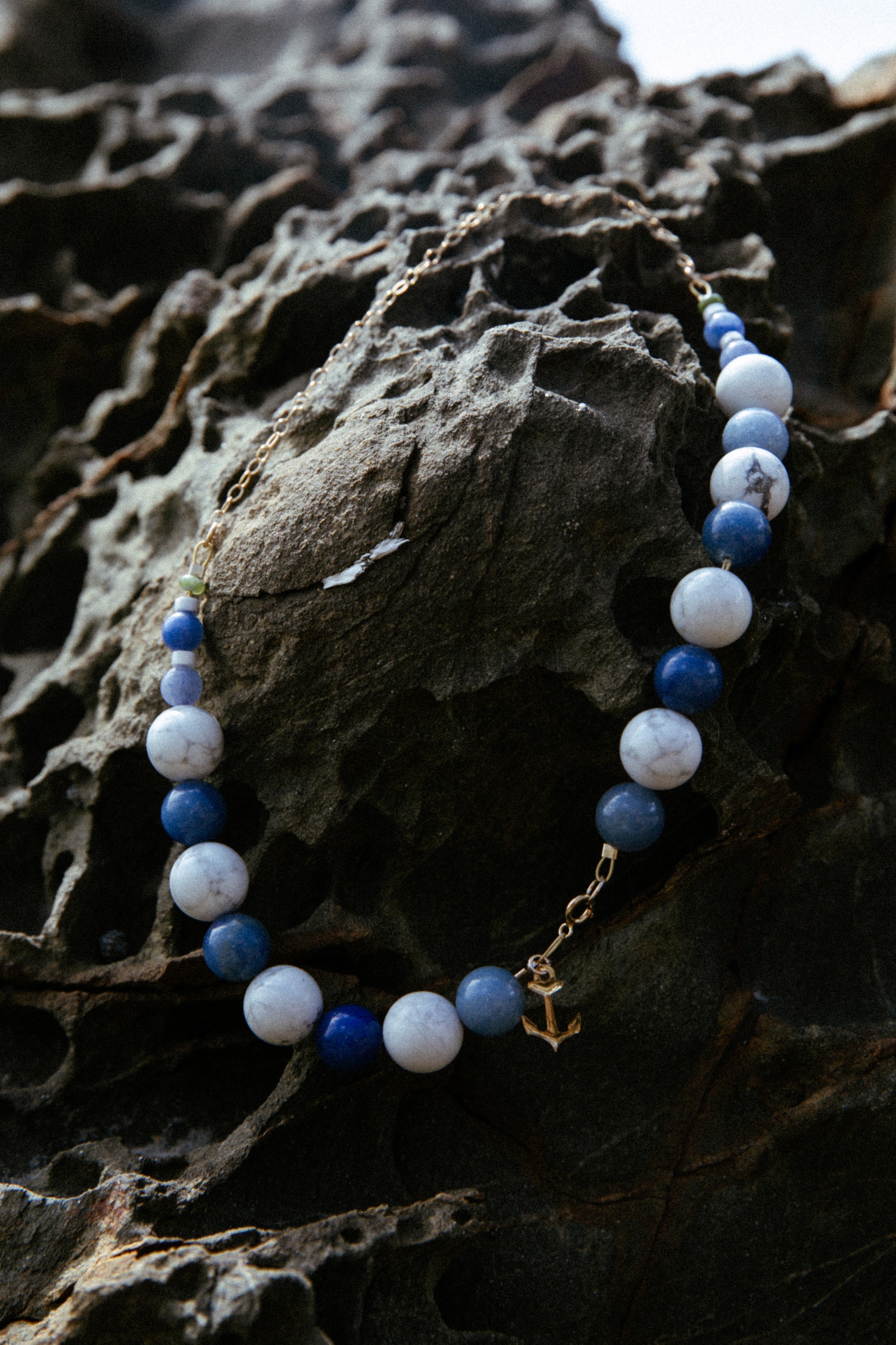A necklace hanging on rocks at the beach. The jewellery is composed of natural howlyte and blue aventurine semi-precious stones, with a gold-filled  anchor charm. 